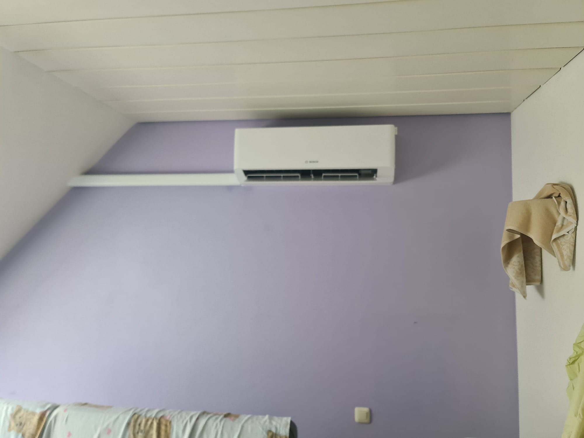 Bosch-Klimagerät an schräger, lavendelfarbener Innenwand unter weißer Decke mit sichtbarem Kabelkanal.